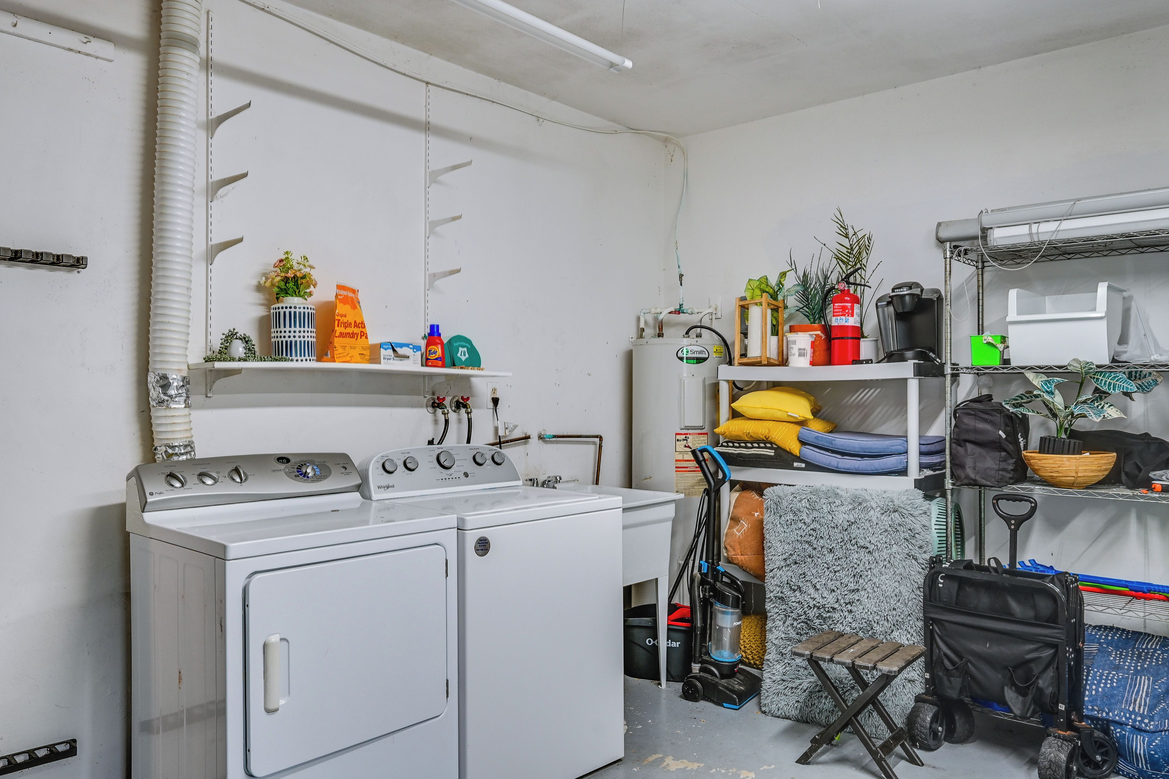 Uma lavanderia com máquina de lavar e secadora, prateleiras com produtos de limpeza, um aspirador e um carrinho com sacos.
