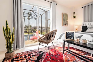 A modern hotel room with a large sliding glass door leading to a pool area, a wicker chair, a bed with a black and white striped bedspread, and a sign with a message. - Pool & Dock! Walk to Downtown Dining & Nightlife (Cape Coral)