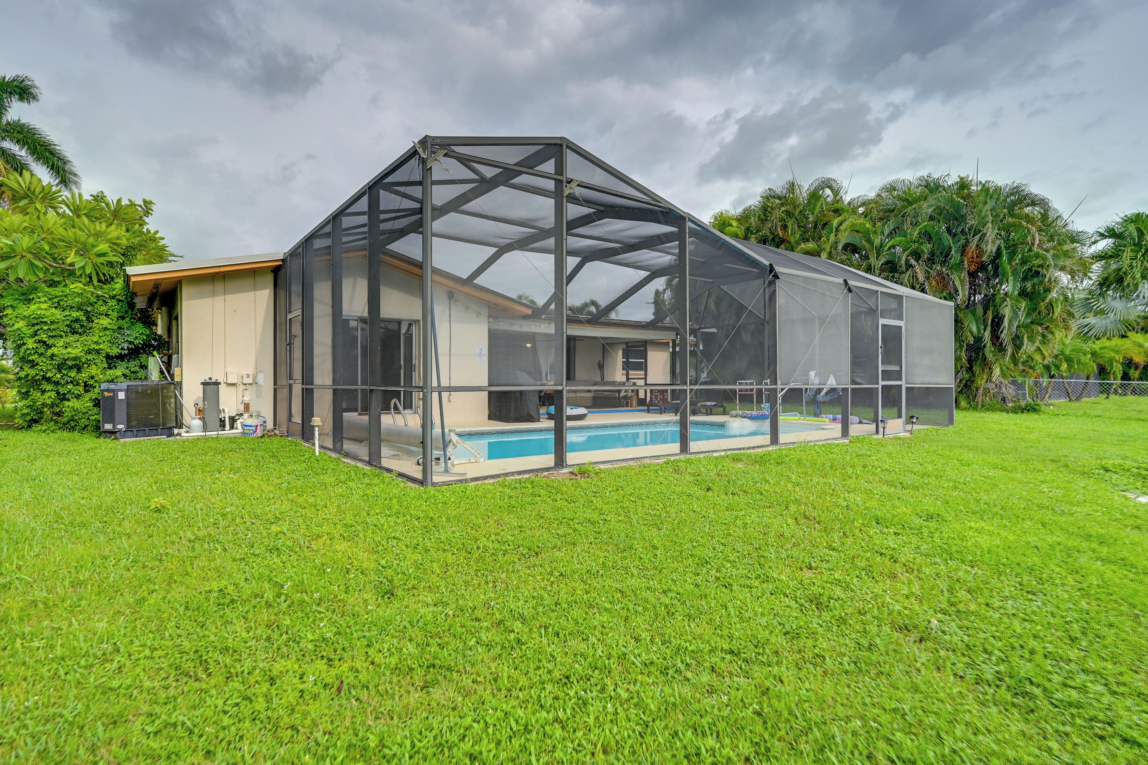 A covered outdoor swimming pool with a glass enclosure, surrounded by a well-maintained lawn and tropical vegetation.