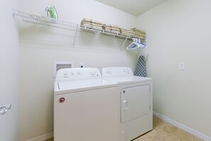 A laundry room with a front-loading washer and dryer, a drying rack with towels, and a shelf with cleaning supplies. - Solterra Resort Home w/ AI Vending & Private Pool! (Davenport)
