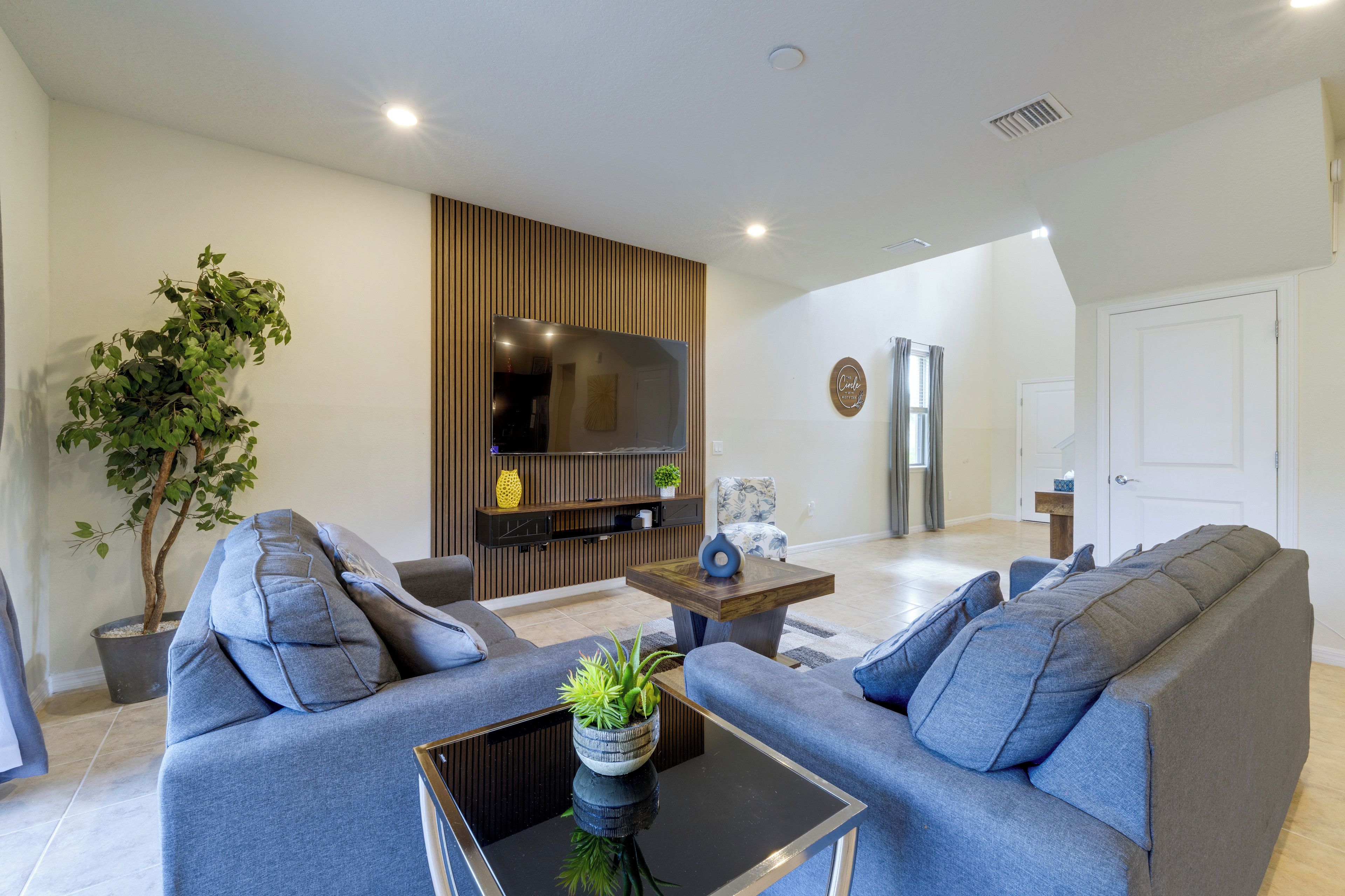 A modern living room with a grey sofa set, a coffee table with a plant, and a flat-screen TV mounted on a wooden panel wall.