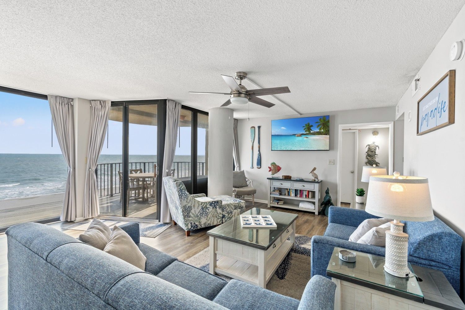 A spacious living area with a balcony overlooking the ocean, a ceiling fan, and a bookshelf.