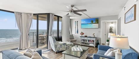A spacious living area with a balcony overlooking the ocean, a ceiling fan, and a bookshelf.