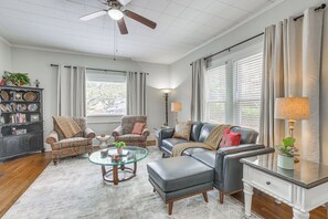 Uma sala de estar com ventilador de teto, dois poltronas, um sofá, uma mesa de centro de vidro e uma mesa lateral com abajur.
