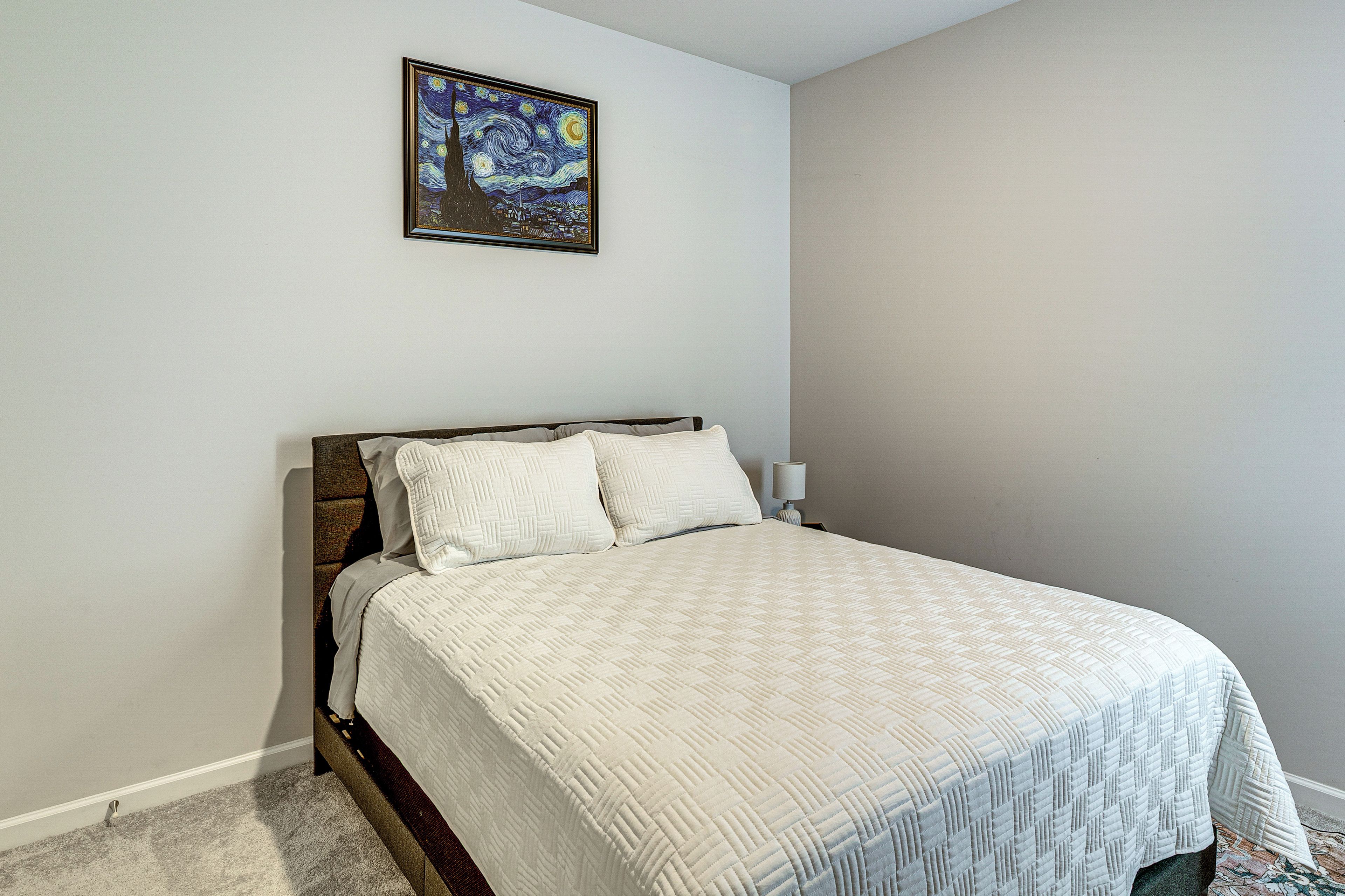 Un dormitorio con cama, mesita de noche, lámpara y un cuadro en la pared.