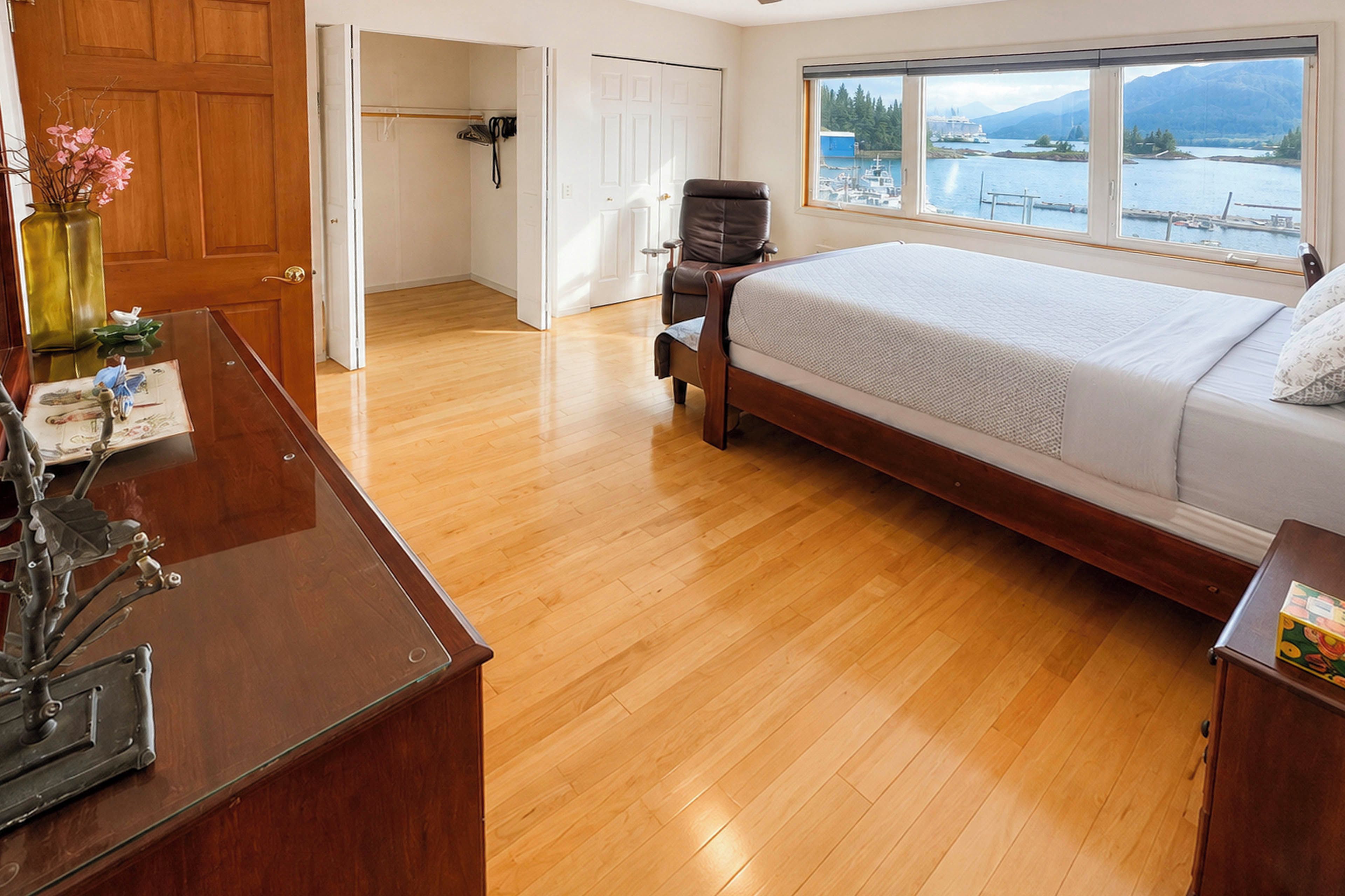 A bedroom with a wooden floor, a bed, a desk, and a window with a view of a body of water and mountains.