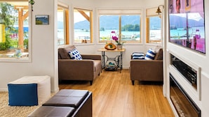 A cozy living room with two sofas, a coffee table, and a fireplace. Large windows offer a view of a lake and mountains.