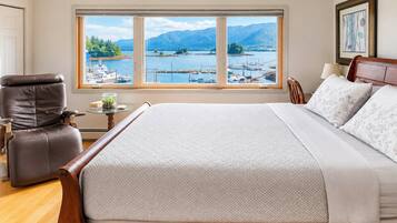 Une chambre avec un grand lit, une chaise, une petite table sur laquelle se trouve un récipient en verre, et une vue sur une marina et des montagnes, visible à travers une grande fenêtre.
