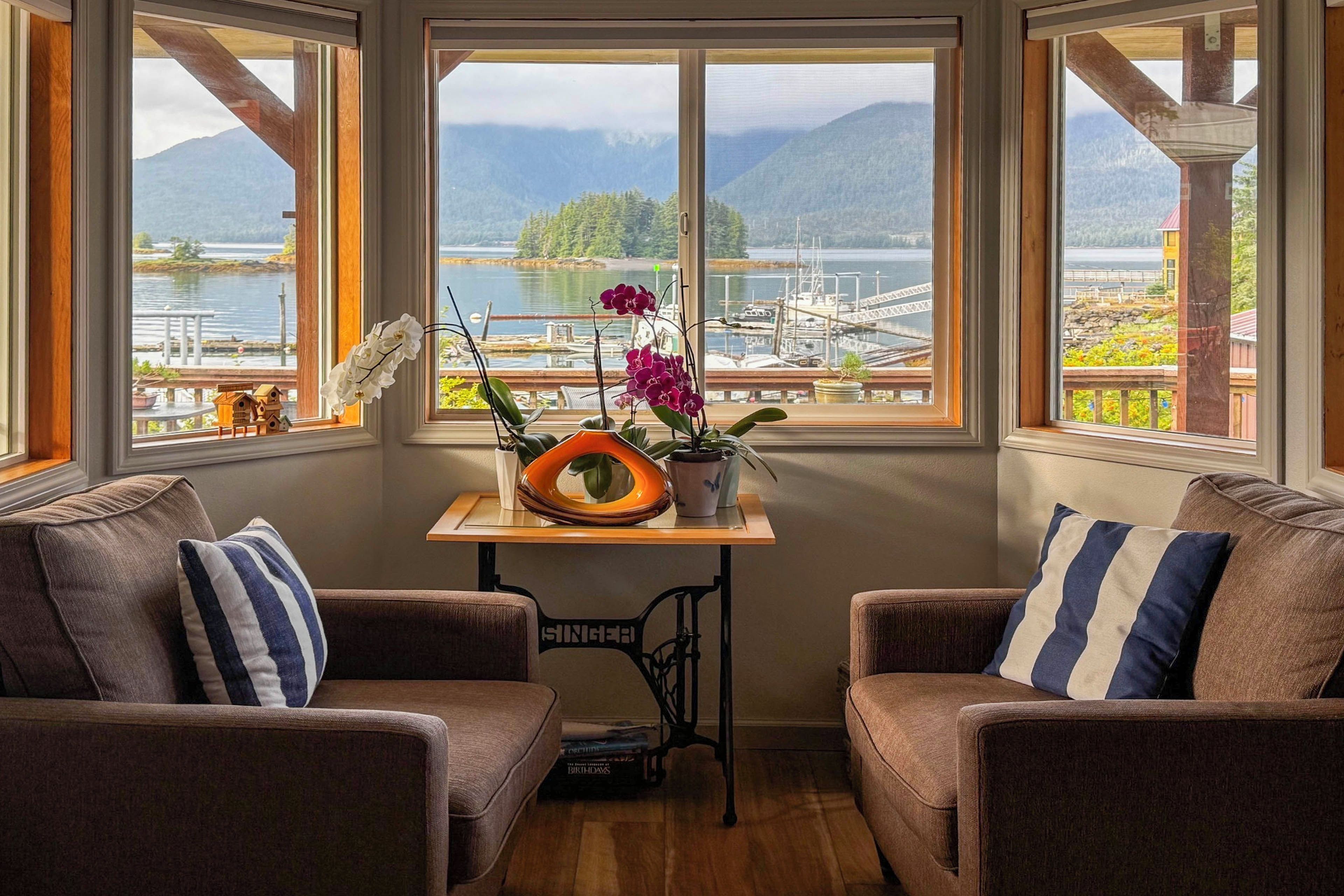 A cozy room with two brown sofas, a small table with potted plants, and a view of a marina and mountains through large windows.