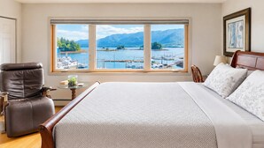 A bedroom with a large bed, a chair, a small table with a glass container, and a view of a marina through a large window.