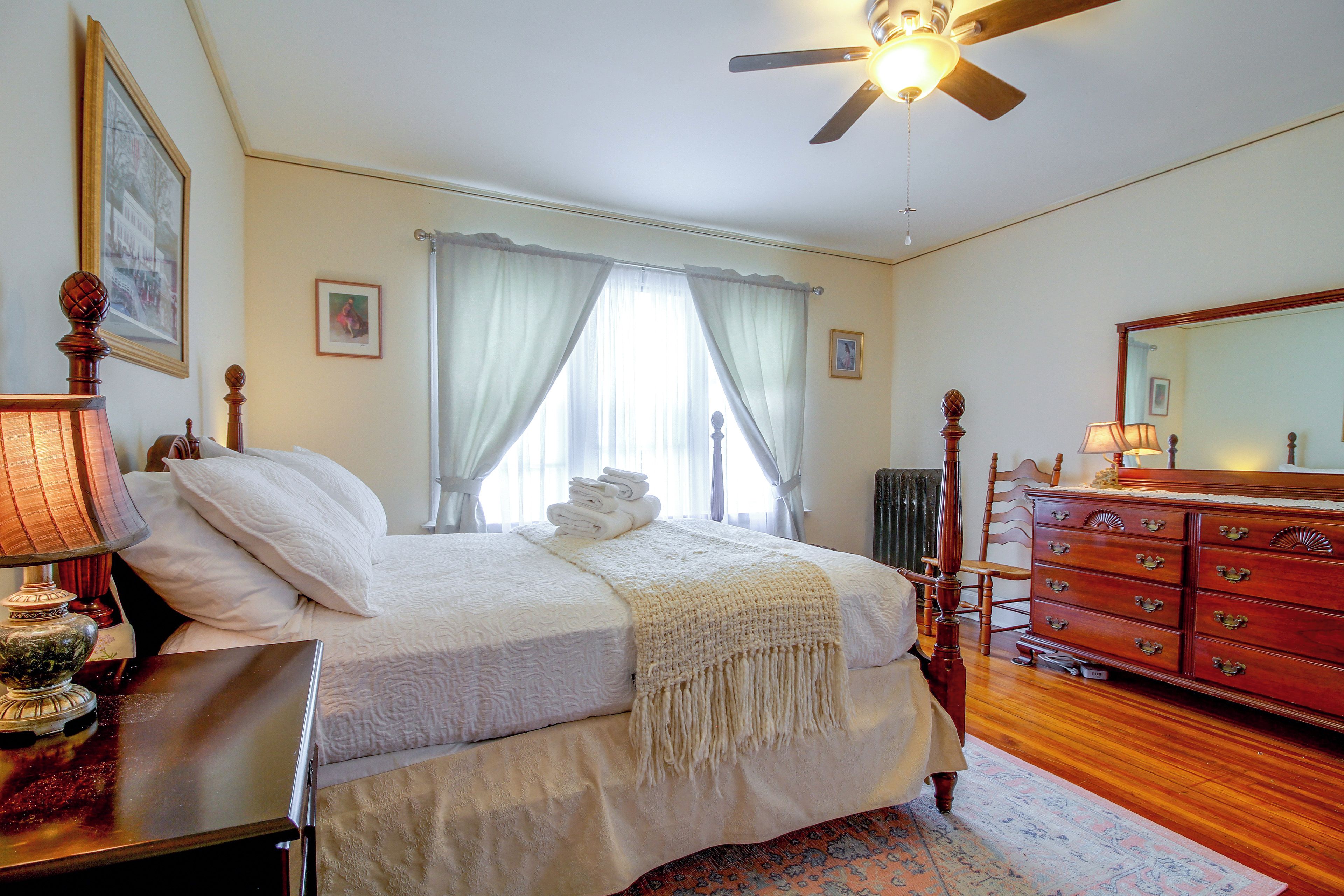 A bedroom with a bed, a dresser, a mirror, a ceiling fan, and a window with curtains.