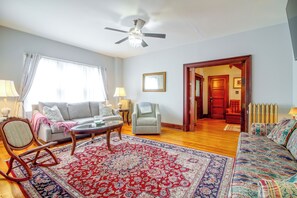 A living room with a ceiling fan, a sofa, a patterned rug, a wooden rocking chair, and a door leading to another room.