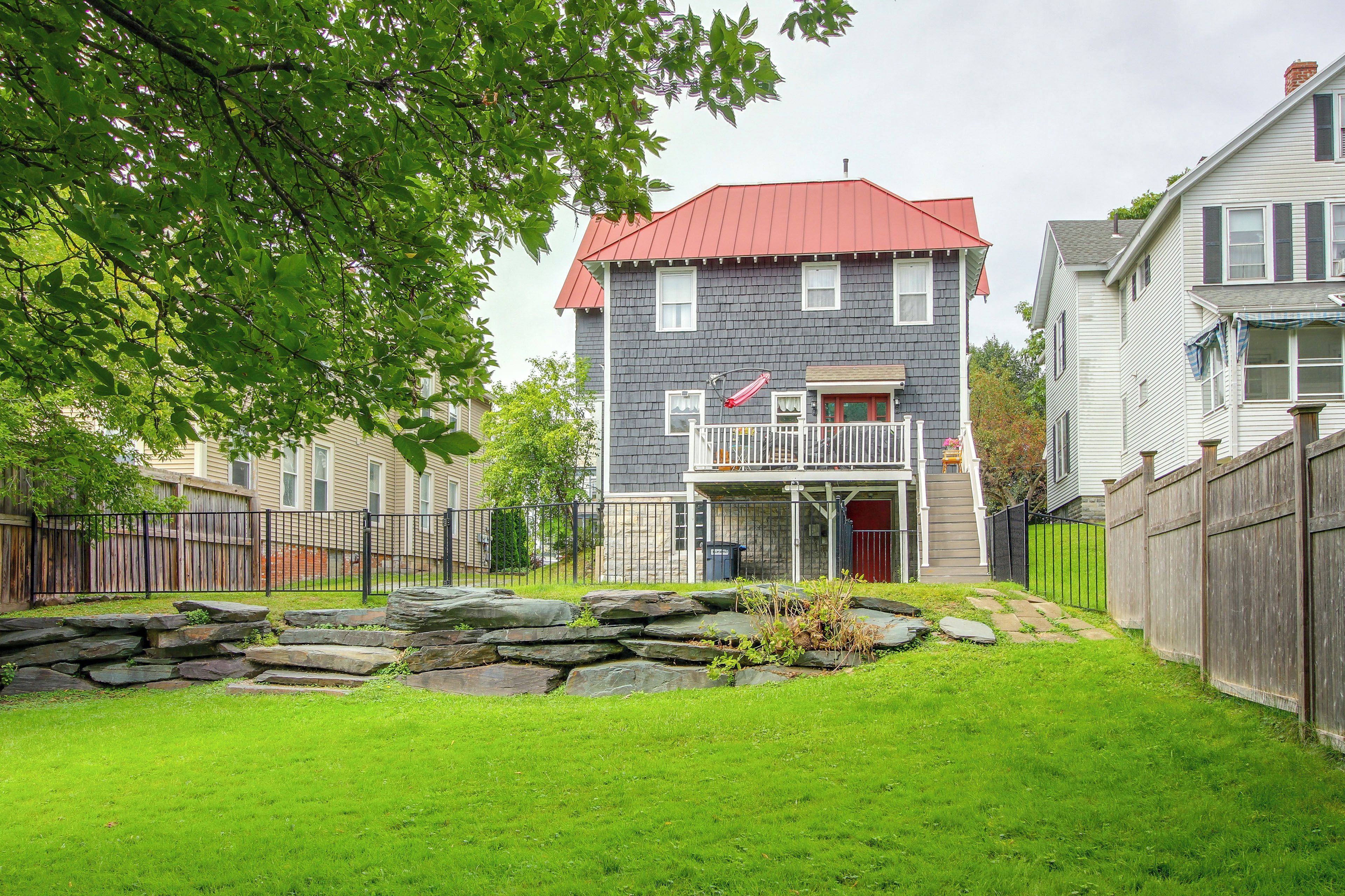 A two-story house with a red roof, a balcony, and a stone wall in the front yard.