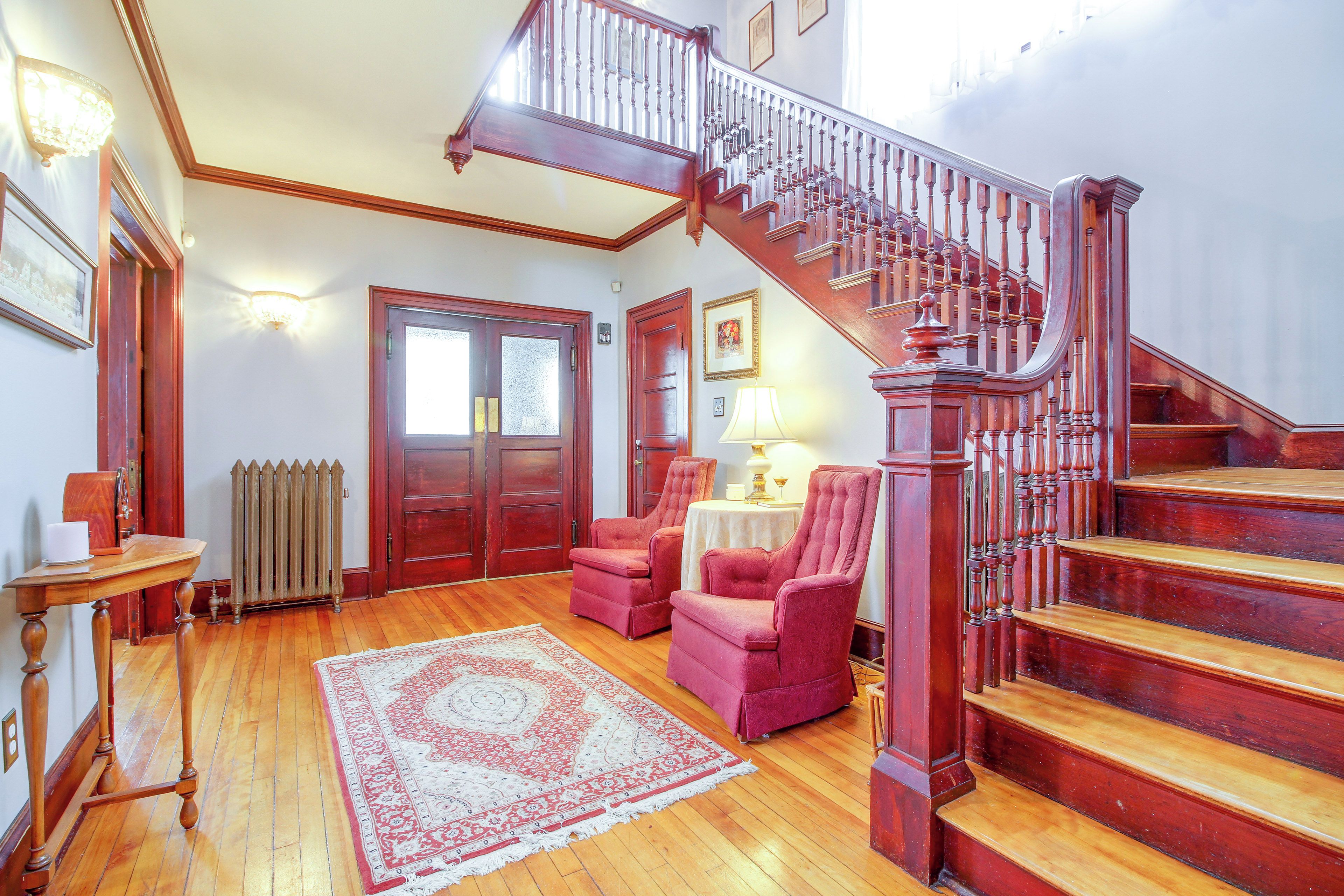 A spacious room with a wooden staircase, a radiator, a table with a lamp, and two armchairs.