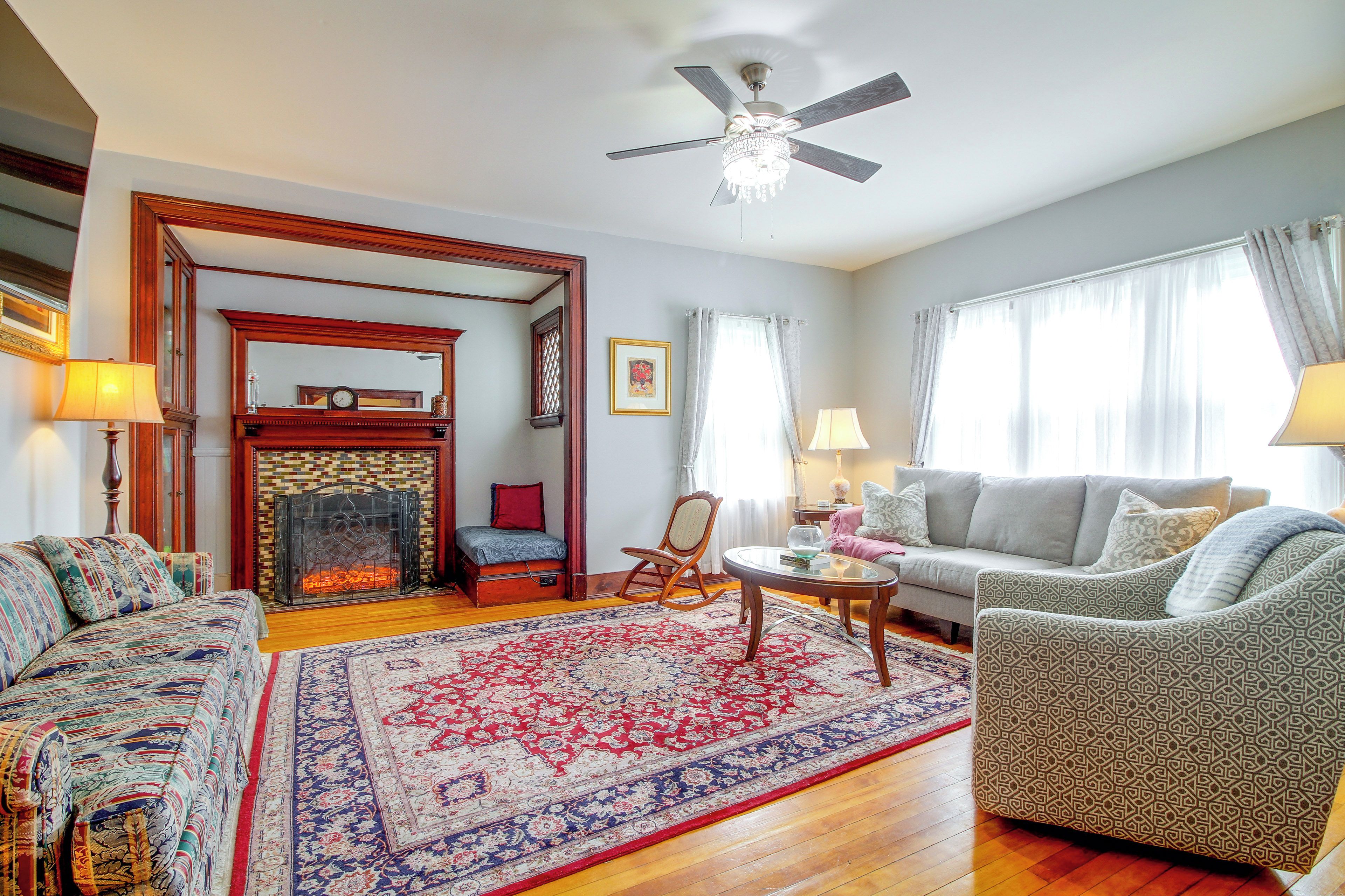 A traditional living room with a fireplace, a ceiling fan, and a large window with curtains.