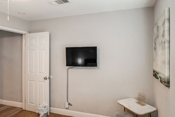 A room with a white door, a mounted flat-screen TV, a small white table with a gold wire shelf, and a painting on the wall.