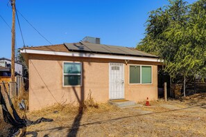 A small, single-story house with a brown exterior, a white door, and a small front yard with dry grass and a red traffic cone.