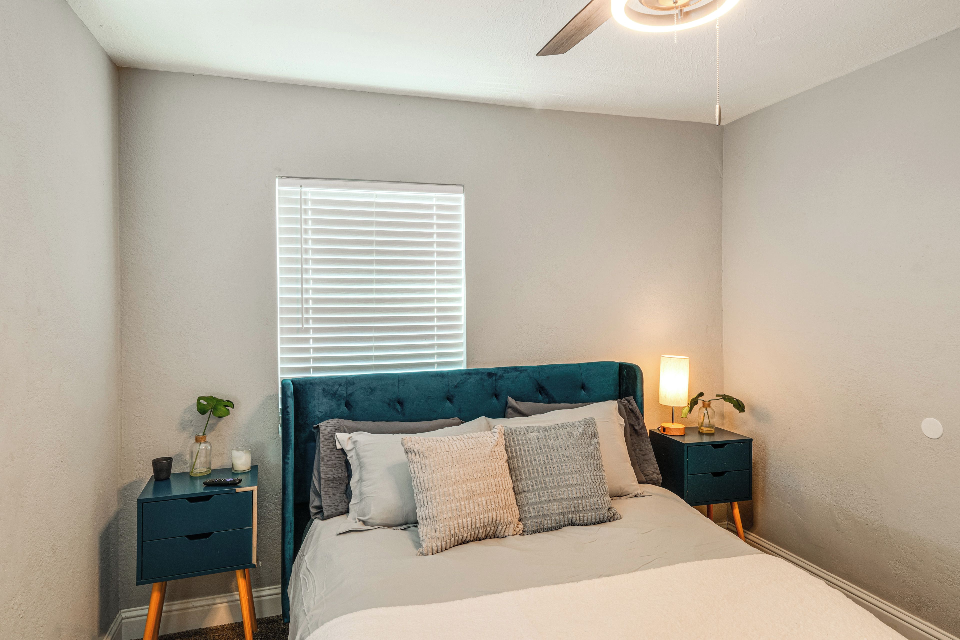 A bedroom with a bed, bedside tables, a window with blinds, a ceiling fan, and a nightstand with a lamp.