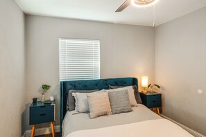 Un dormitorio con cama, mesitas de noche, una ventana con persianas, un ventilador de techo y una mesita de noche con lámpara.