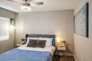 A bedroom with a bed, a ceiling fan, a nightstand with a lamp, and a wall painting.