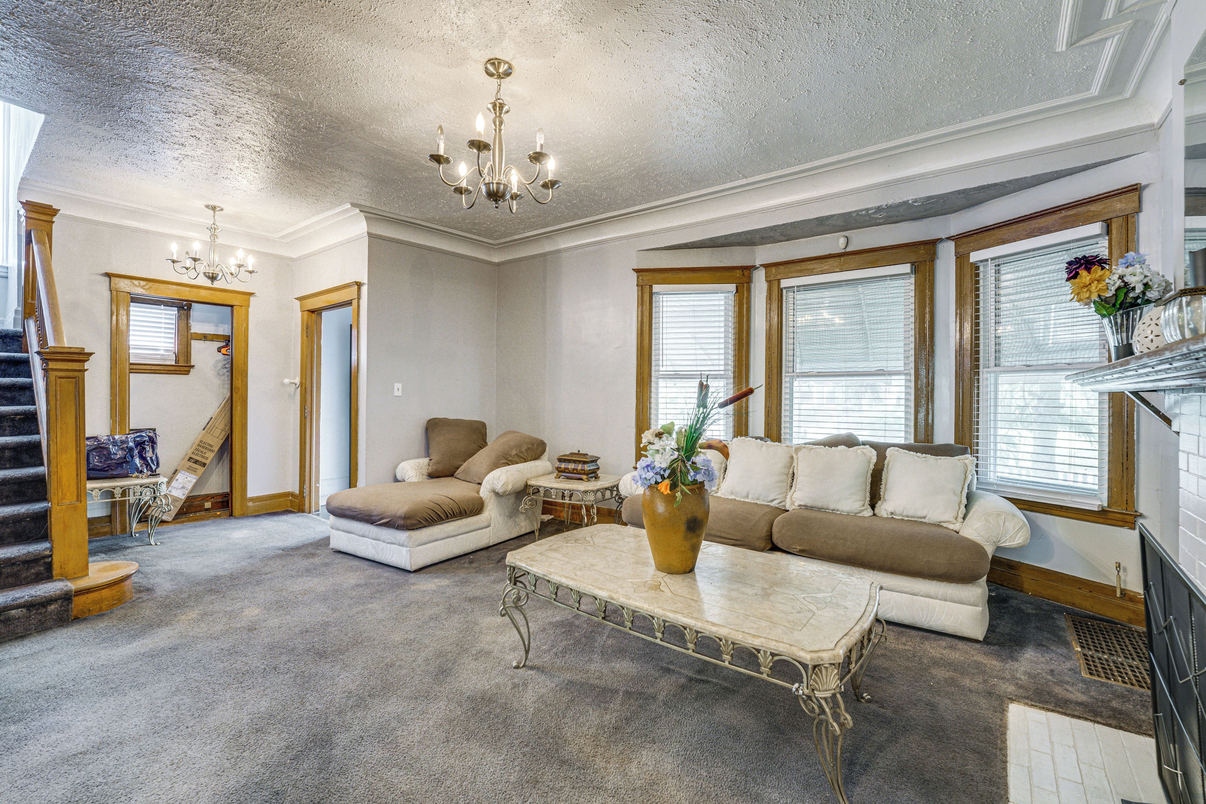 A spacious living room with a high ceiling, a chandelier, a fireplace, and a large window with blinds.