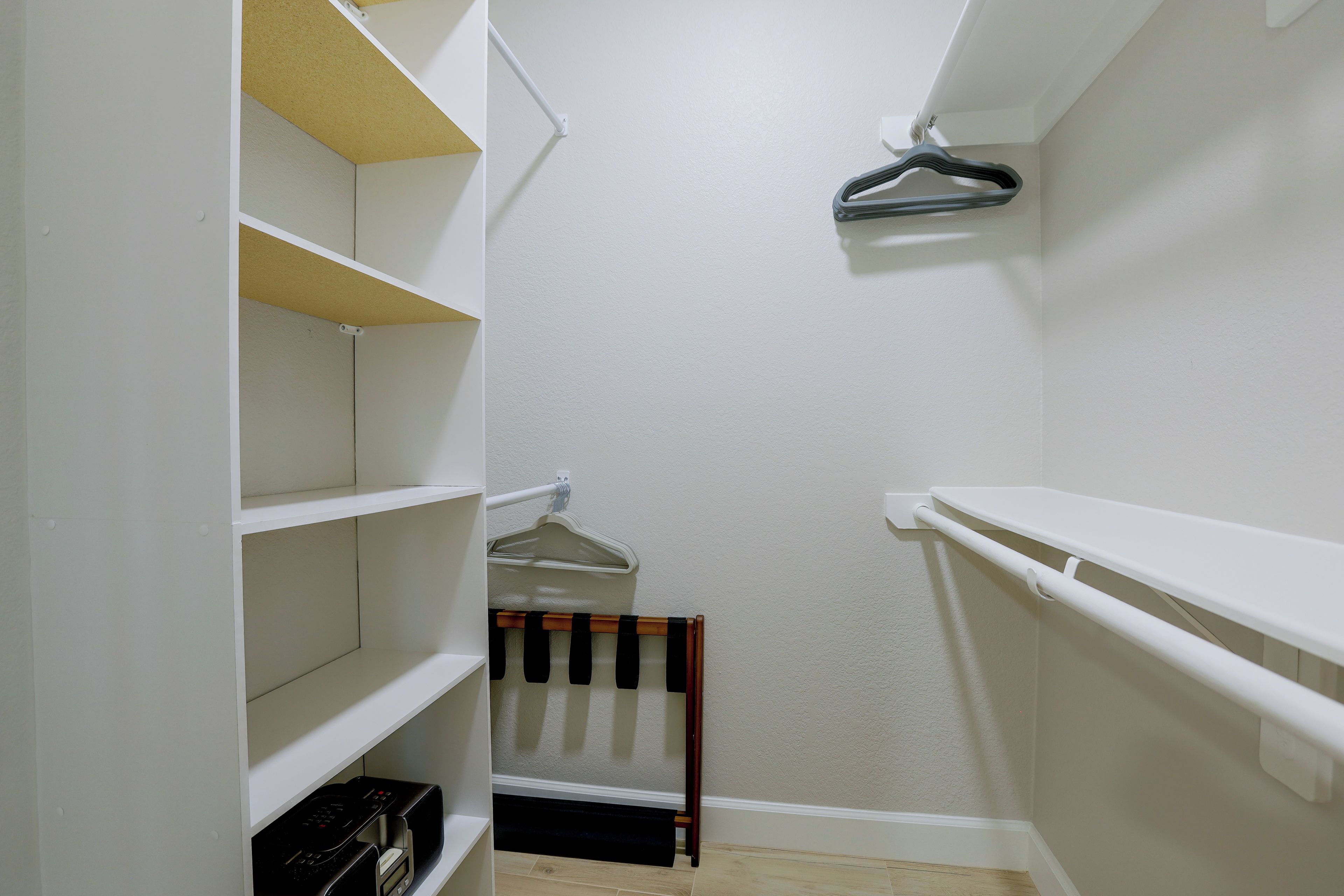 A narrow walk-in closet with white shelving, a hanging rod, and a wooden bench.