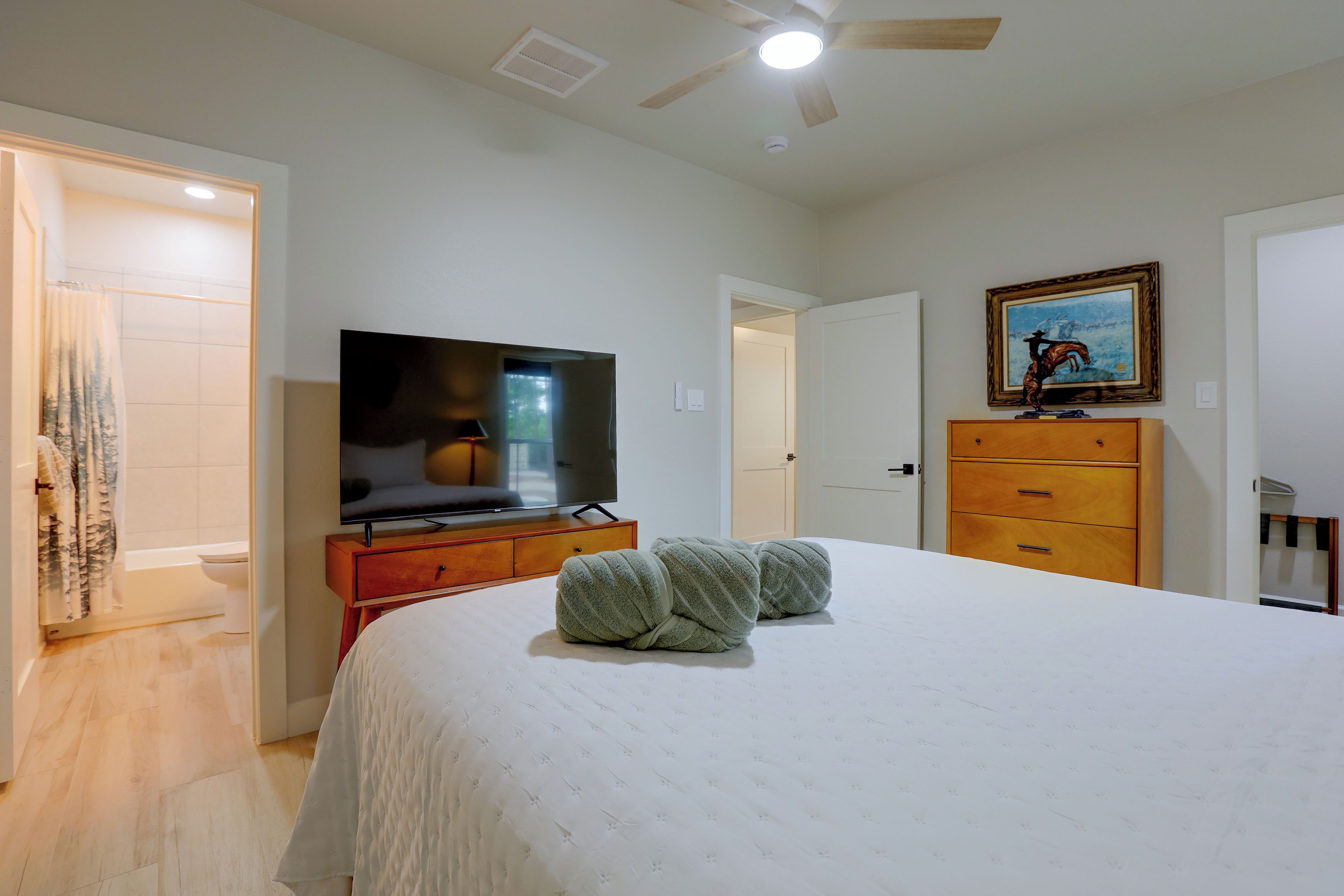Un dormitorio con cama, televisor sobre un aparador de madera, un cuadro en la pared y un ventilador de techo.