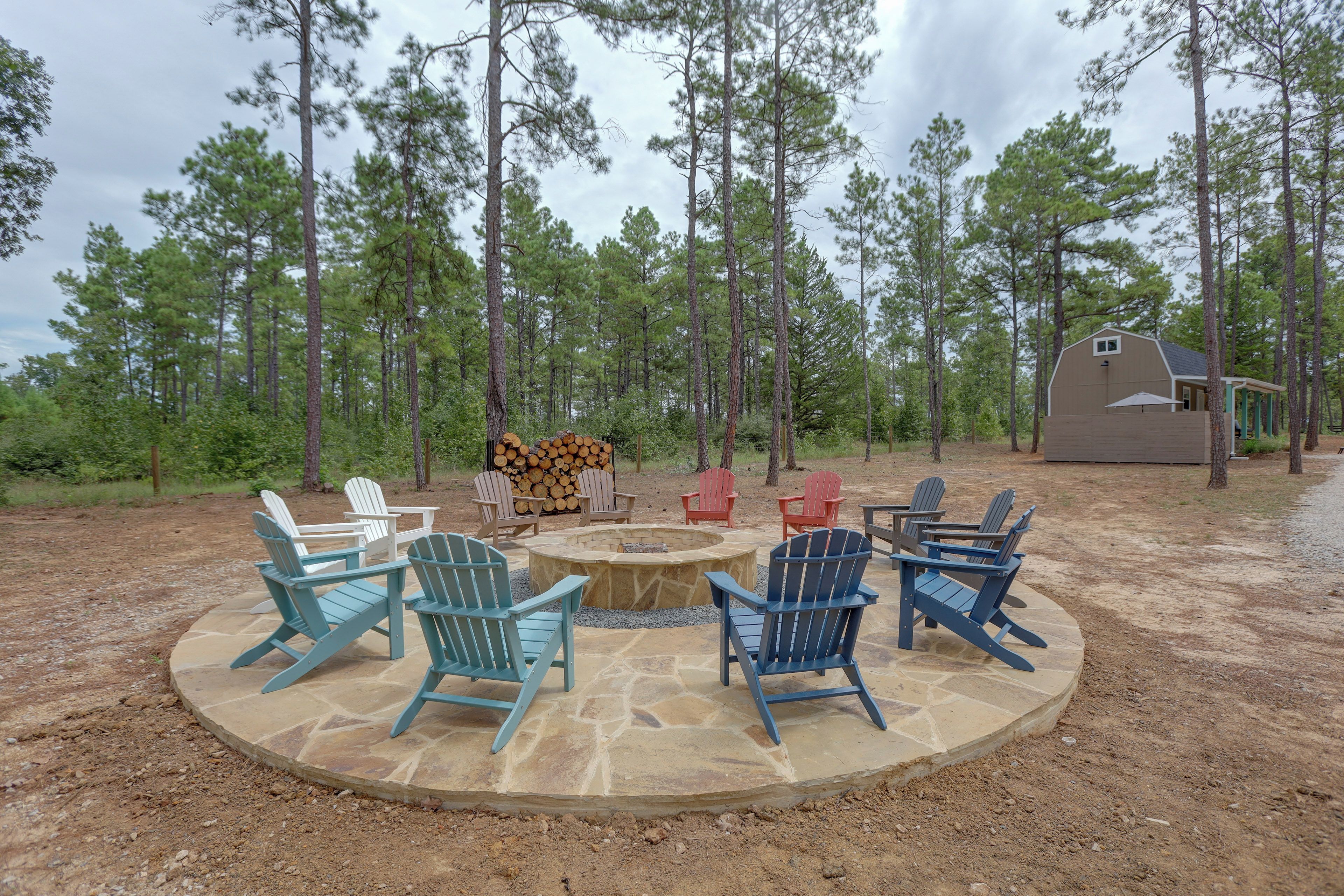 A circular outdoor seating area with Adirondack chairs and a fire pit, surrounded by a forested landscape.