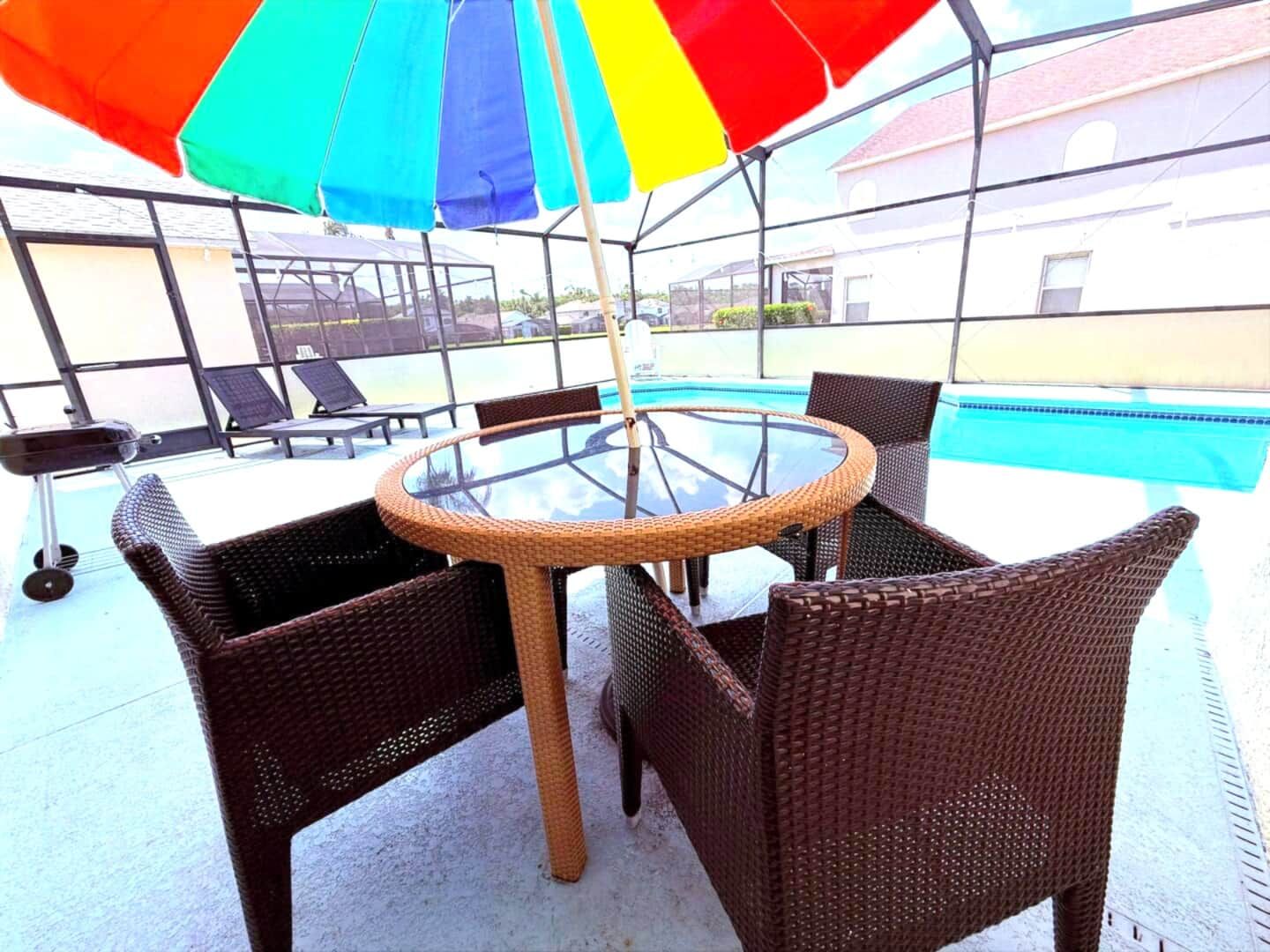 Una mesa con cubierta de vidrio y sillas de mimbre, bajo un paraguas de varios colores, con una piscina al fondo.