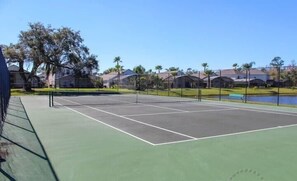 A tennis court with a net, surrounded by a fence, and a body of water in the background. - Ref 301. 3 Bedroom villa own pool. Lake Berkley Resort. (Osceola County)