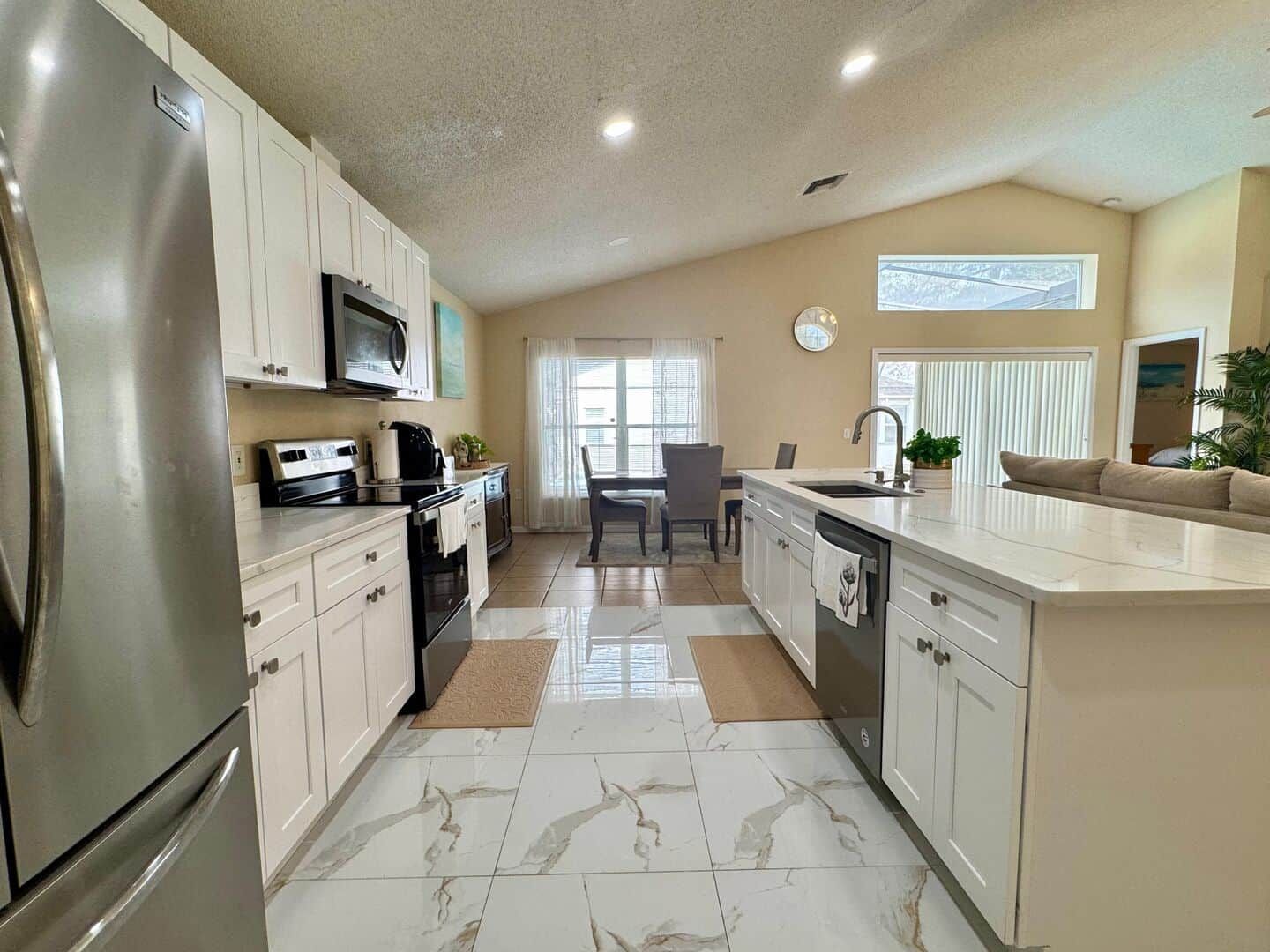 A modern kitchen with a refrigerator, microwave, stove, and sink. There is a dining area with a table and chairs, and a window with blinds.