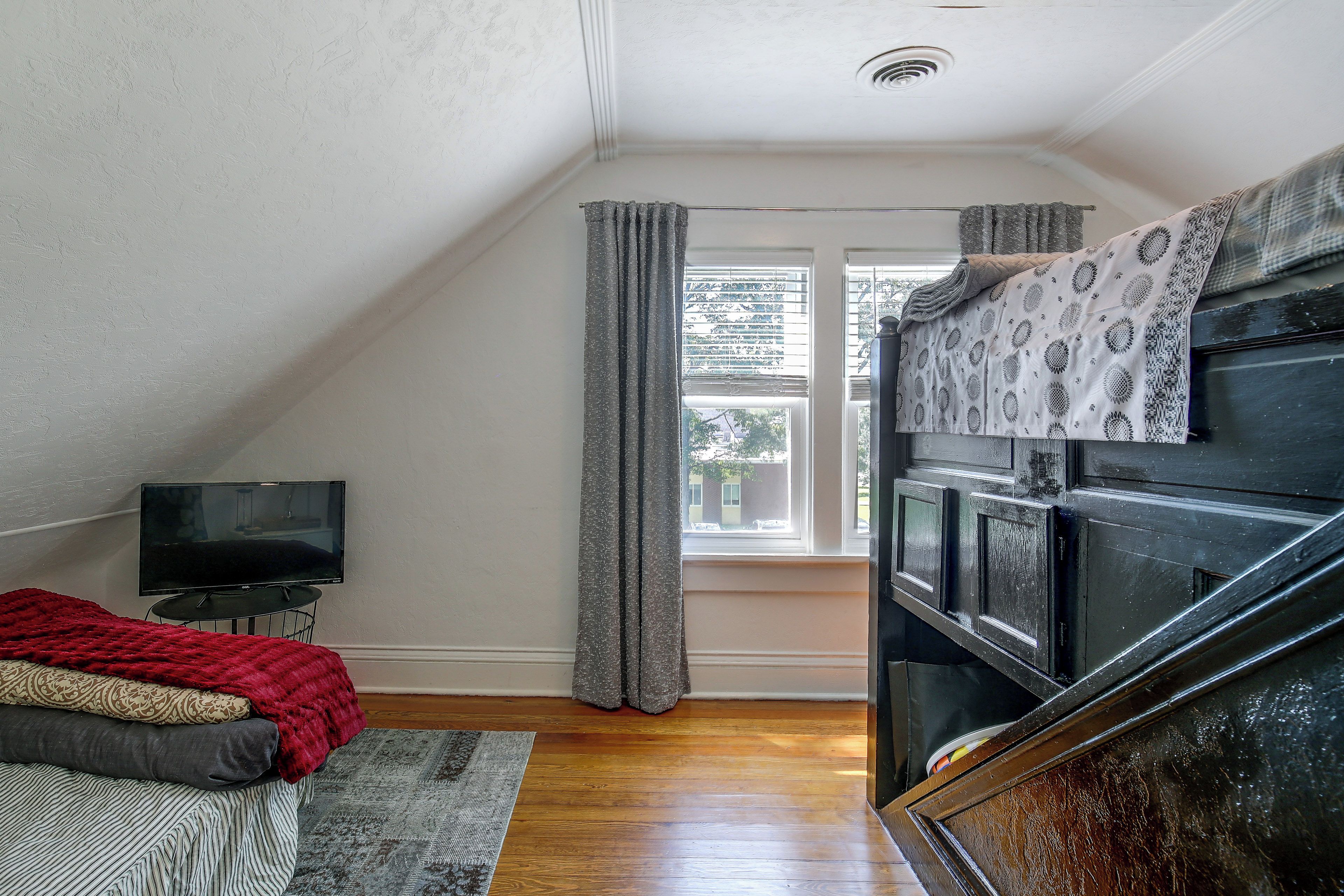 A bedroom with a TV, a bed with a red blanket, a window with curtains, and a staircase.