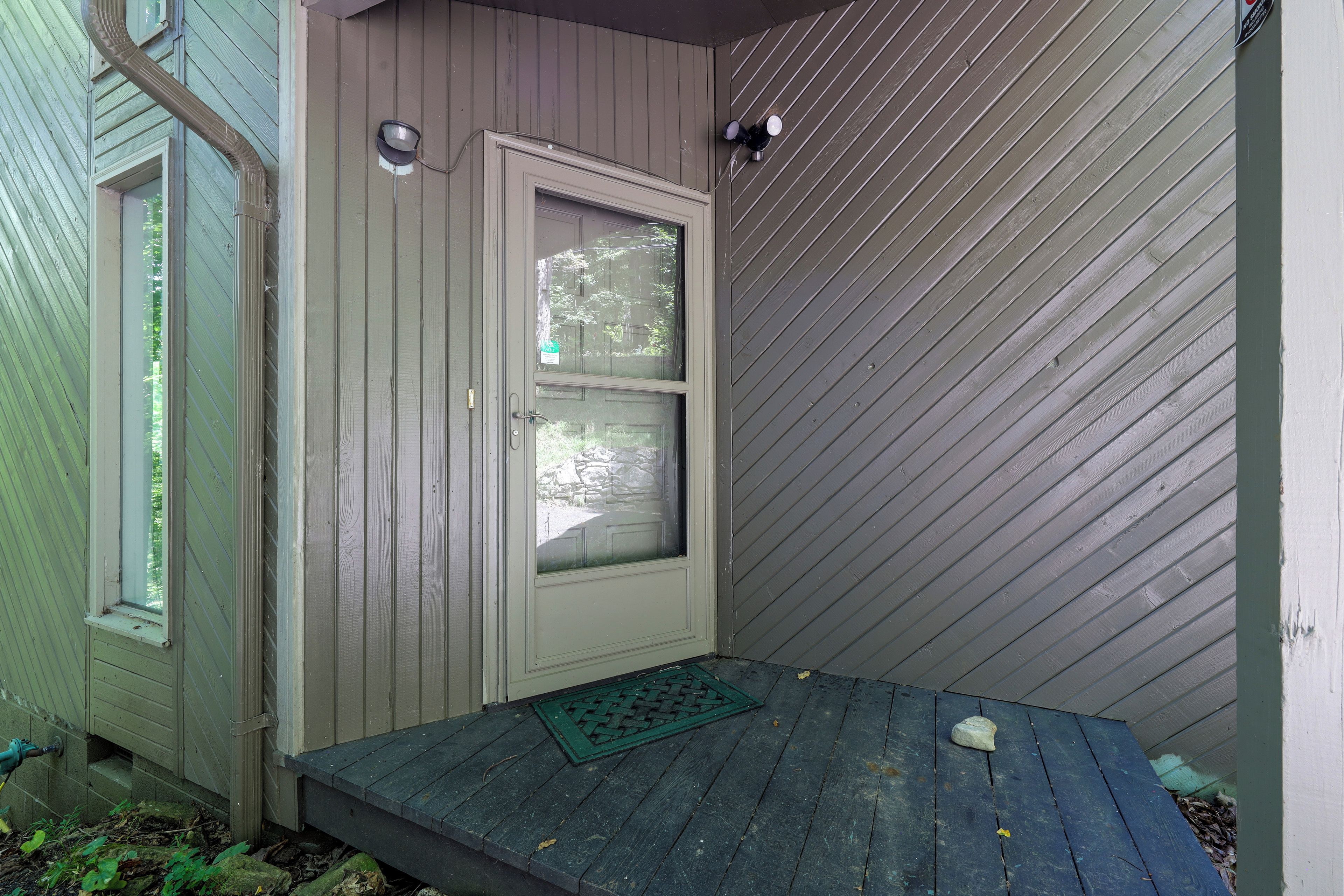 Una porta di legno con una finestra, un piccolo portico con un tappetino verde e un muro con rivestimento a diagonale.