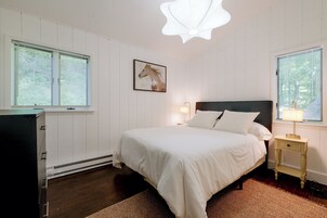 Quarto com cama, mesas de cabeceira, luminária, um quadro na parede e janelas com persianas.