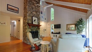 Una sala de estar con chimenea de piedra, muebles blancos, un televisor y un ventanal grande.