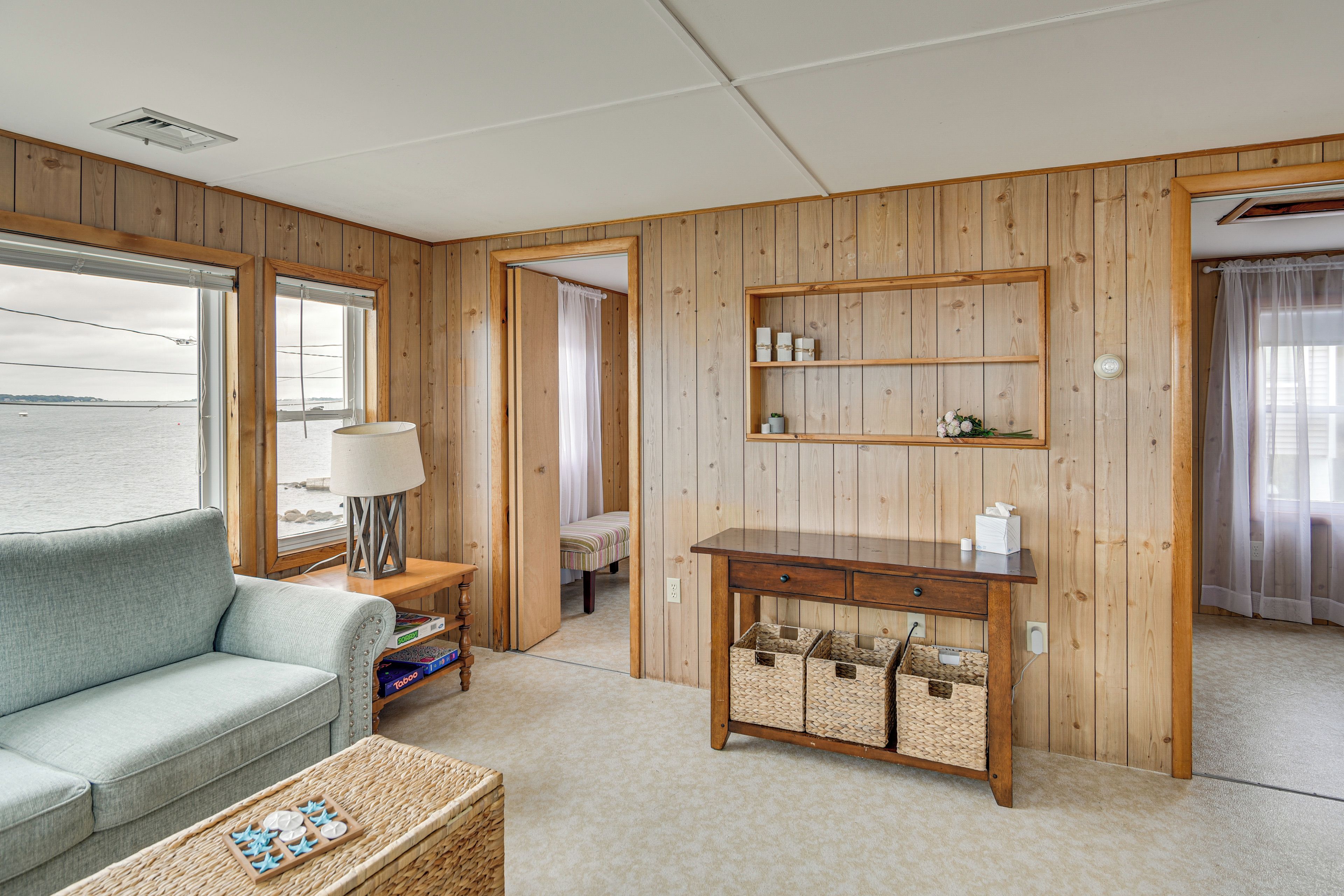 Una habitación con un panel de madera, un sofá, un escritorio con cestas de almacenamiento y vista al mar a través de una ventana.