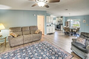 Un salon avec un canapé, une table basse et un tapis. On y trouve également un coin repas avec une table et des chaises. La cuisine est visible au fond.
