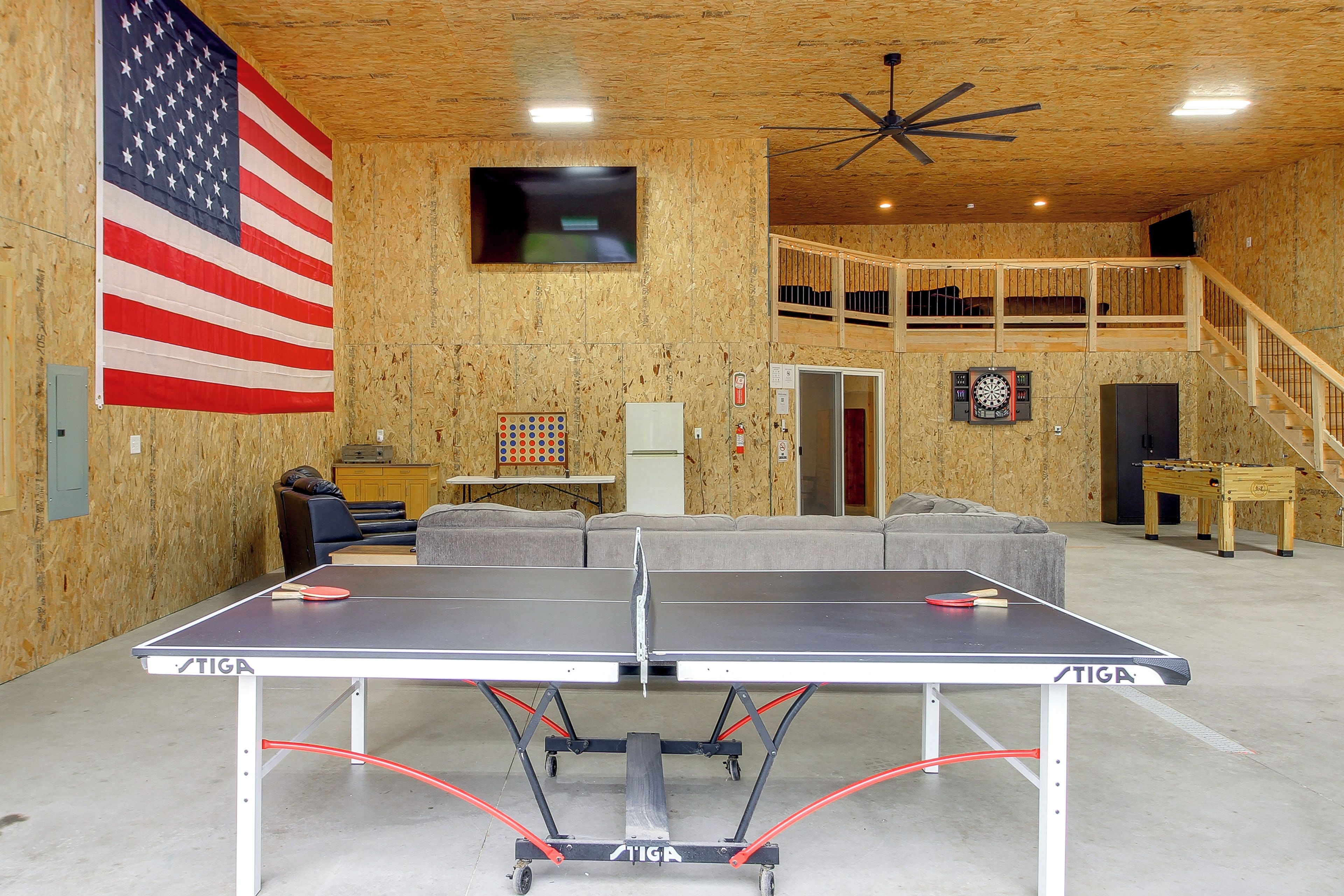 A ping pong table in a room with wooden walls, an American flag, a television, and a dartboard.