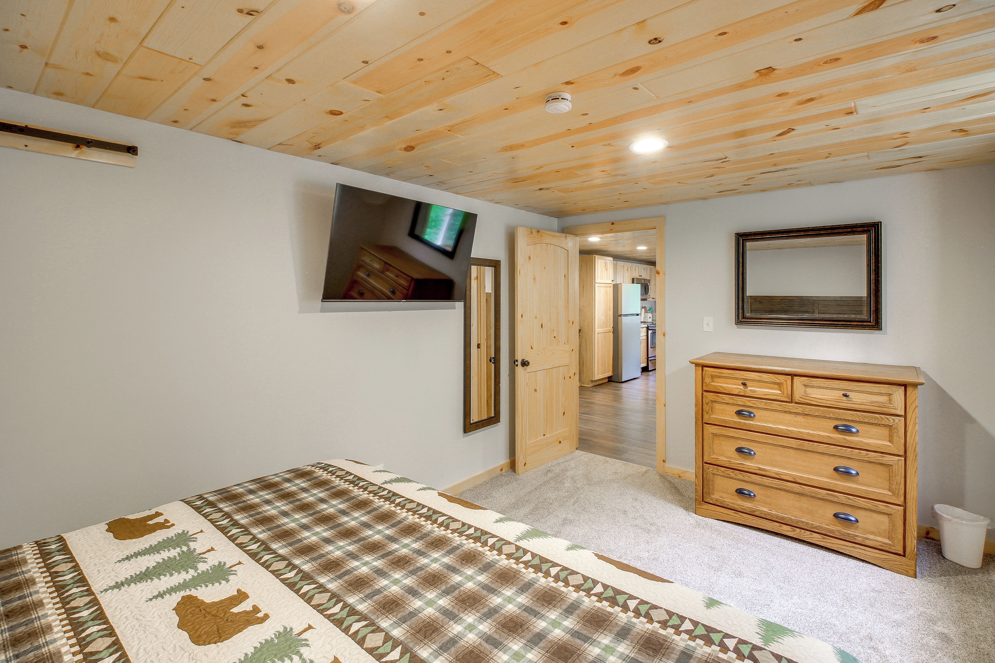 A bedroom with a wooden ceiling, a bed with a patterned bedspread, a dresser, a television, and a door leading to another room.