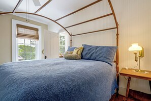 A bedroom with a four-poster bed, a bedside table with a lamp, and a window with blinds.