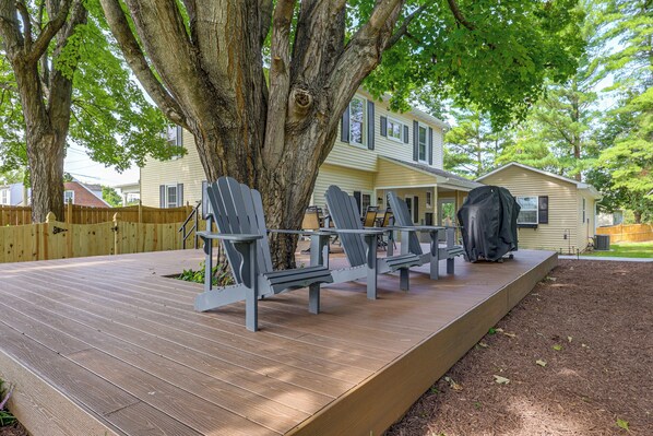 A deck with Adirondack chairs, a covered grill, and a house in the background.