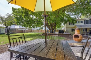Un patio al aire libre con una mesa, sillas, un paraguas amarillo y un árbol grande que da sombra.