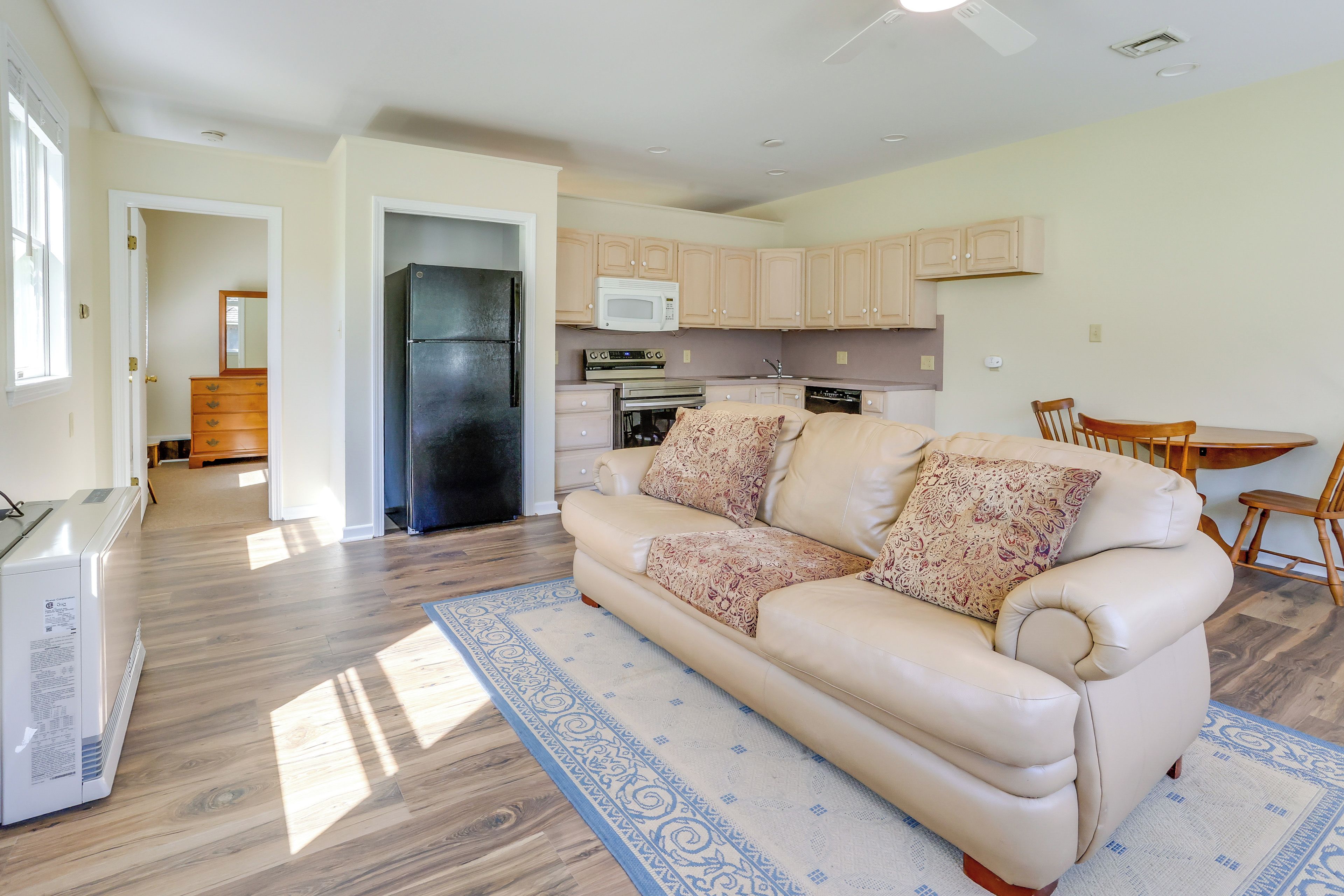 A living room with a beige sofa, patterned cushions, a wooden floor, and a kitchen in the background.