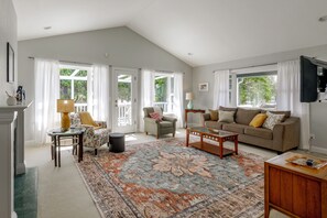 A spacious living room with a high ceiling, a large window, a sofa set, a coffee table, and a television mounted on the wall.
