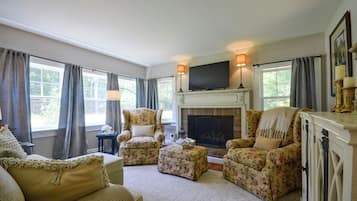A living room with a fireplace, floral armchairs, a television, and large windows with curtains.