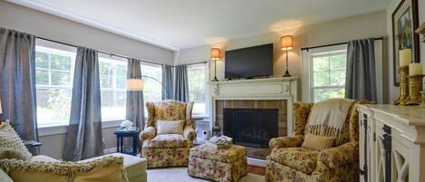 A living room with a fireplace, floral armchairs, a television, and large windows with curtains.