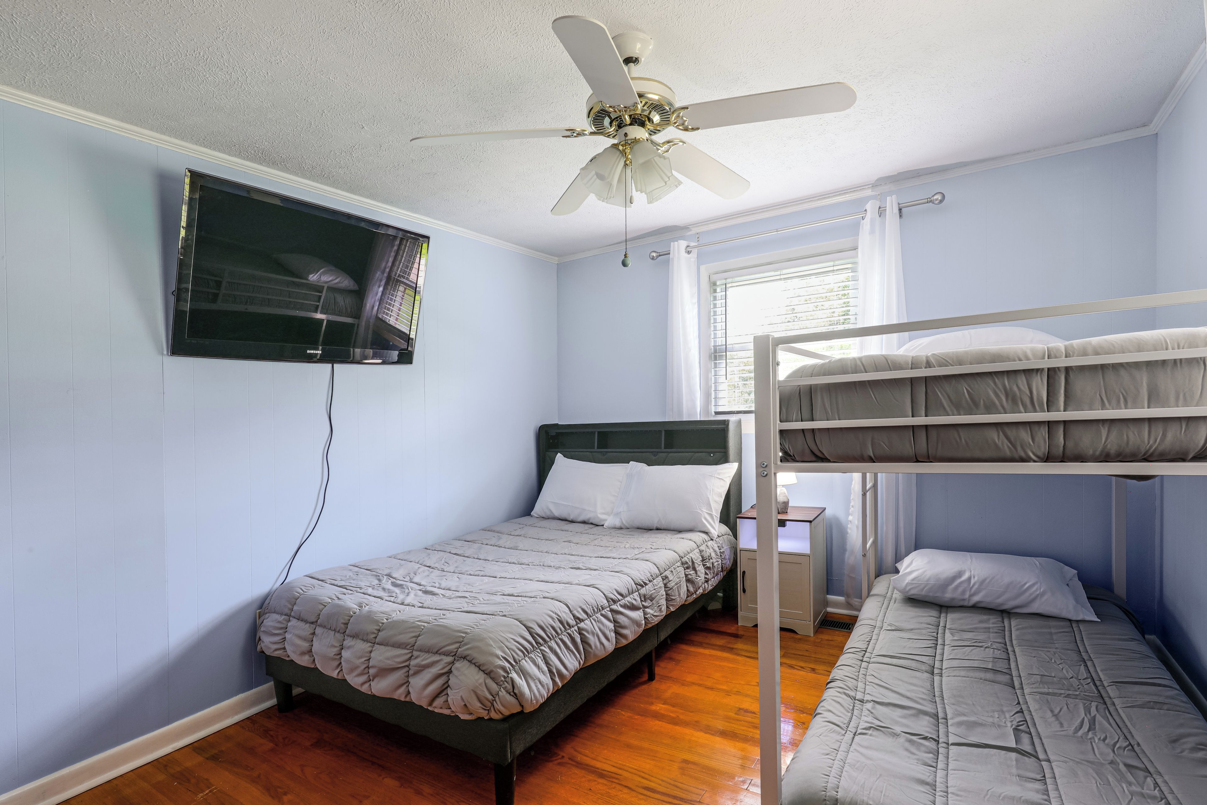 Un dormitorio con una cama individual, literas, un ventilador de techo, una ventana con persianas y un televisor montado en la pared.