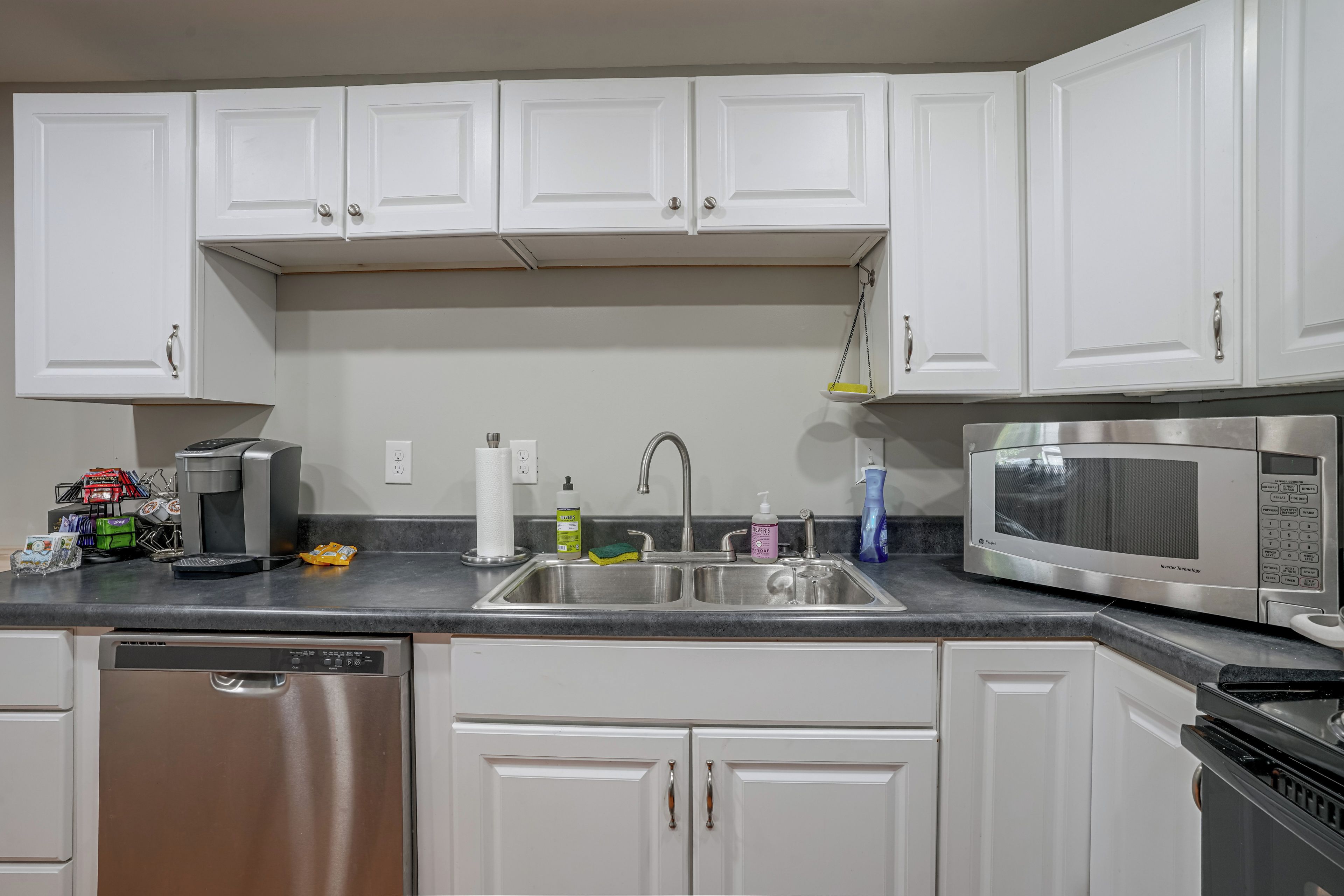 A kitchen with white cabinets, a stainless steel sink, a dishwasher, a microwave, and various kitchen items on the counter.