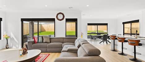 A modern living room with a sectional sofa, a dining area with bar stools, and large windows offering a view of a backyard.