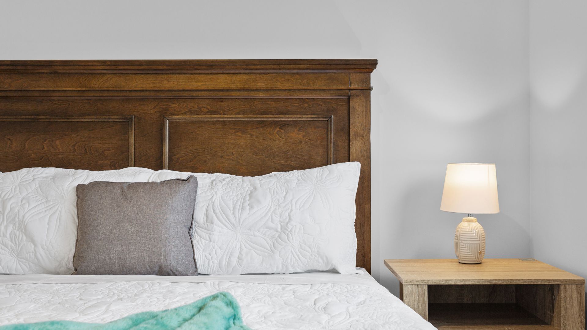 Ein Bett mit Holz-Kopfteil, weißen und grauen Kissen sowie einer Leinen-Decke in Türkis, und ein Nachttisch mit Lampe.